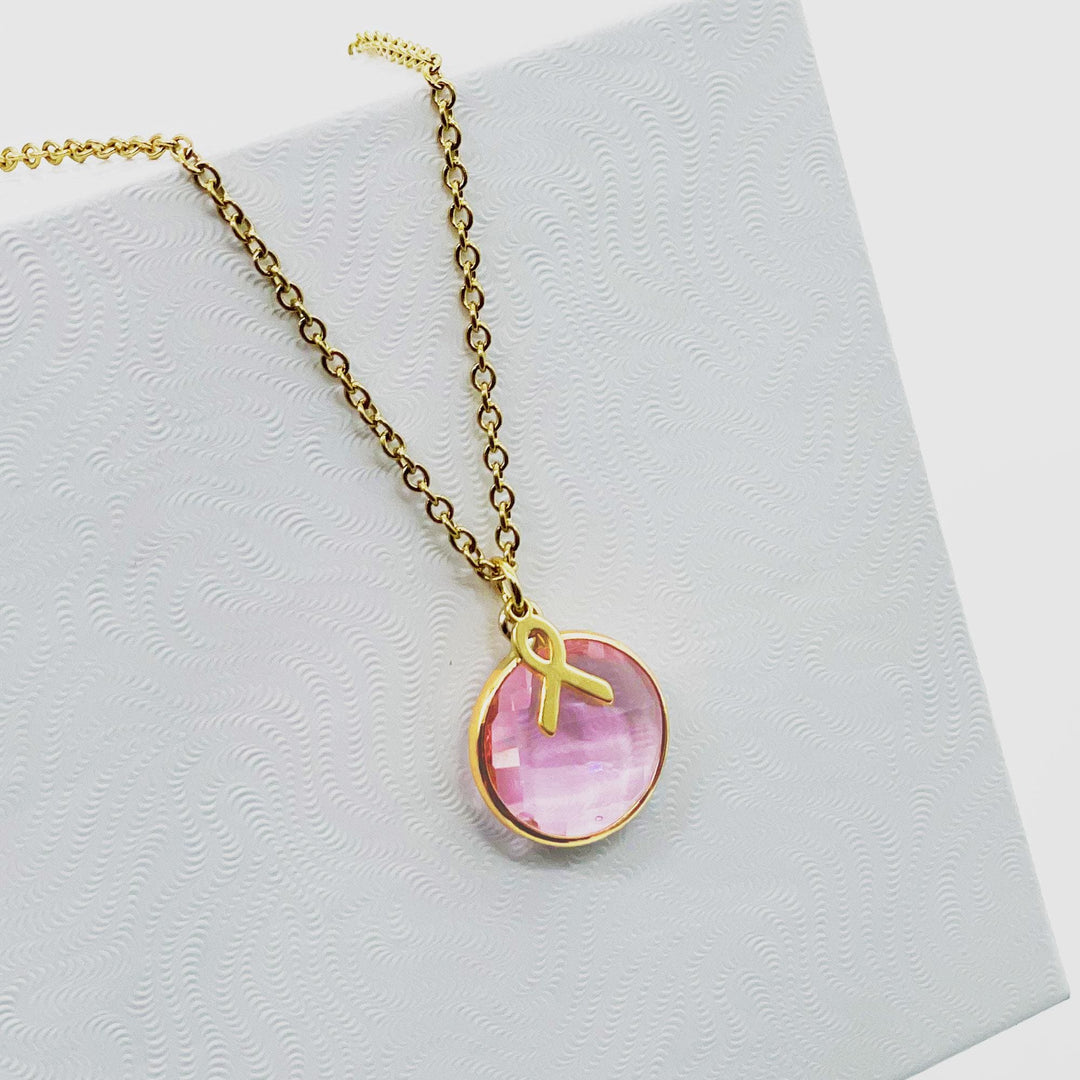 A gold necklace with a pink gemstone pendant is displayed against a white background.