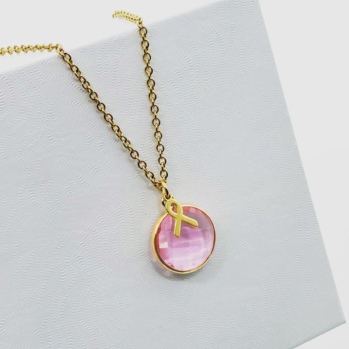 A gold necklace with a pink gemstone pendant is displayed against a white background.