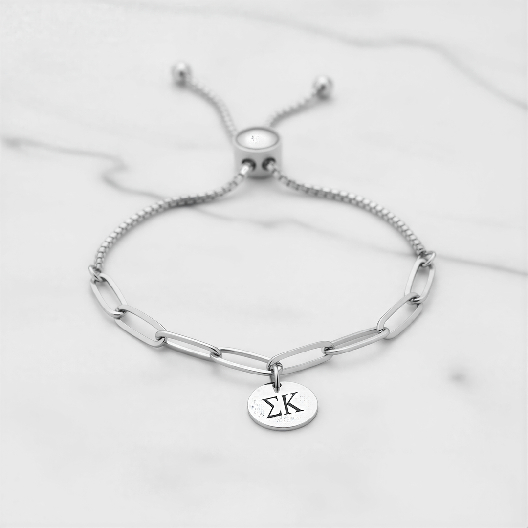 A silver adjustable bracelet with a charm featuring the Greek letter 'Σ' ( Sigma ) on it, placed on a marble surface.