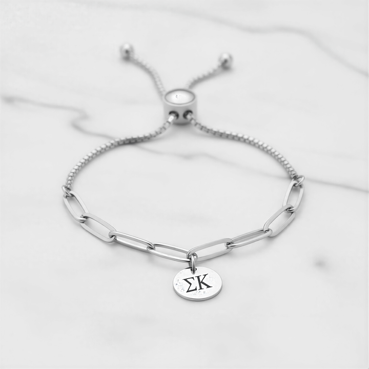 A silver adjustable bracelet with a charm featuring the Greek letter 'Σ' ( Sigma ) on it, placed on a marble surface.