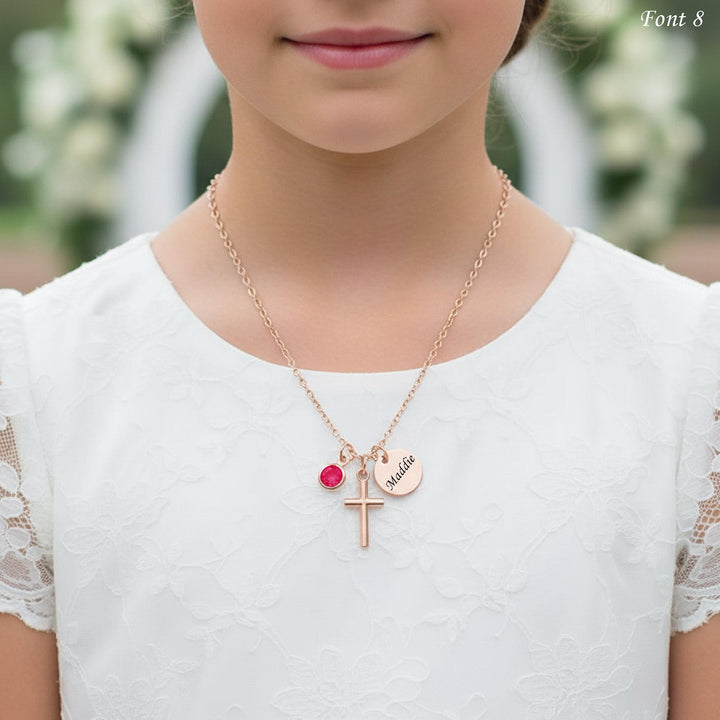 A close-up of a young girl wearing a white lace dress and a gold necklace with a cross pendant and a small red gemstone.
