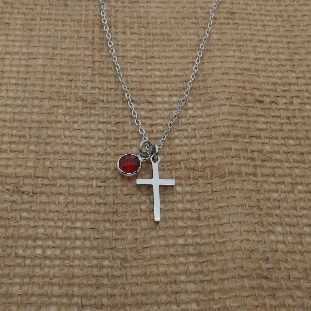 A silver cross necklace with a red gemstone charm is displayed against a woven fabric background.