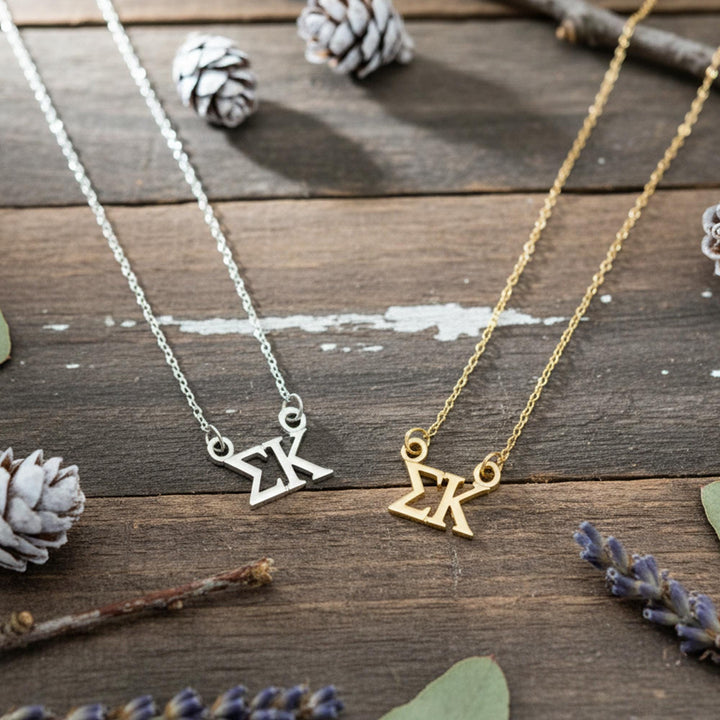 two necklaces with the letters "K" on them, placed on a wooden surface alongside pine cones and other natural elements.