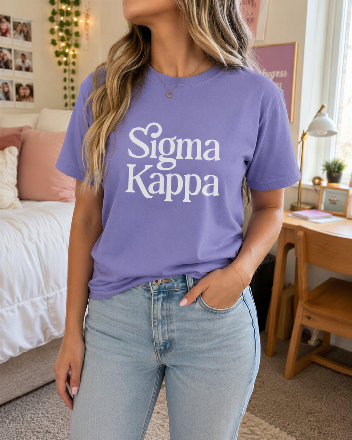 A woman wearing a purple t-shirt with the Sigma Kappa logo stands in a room with a bed, desk, and window in the background.