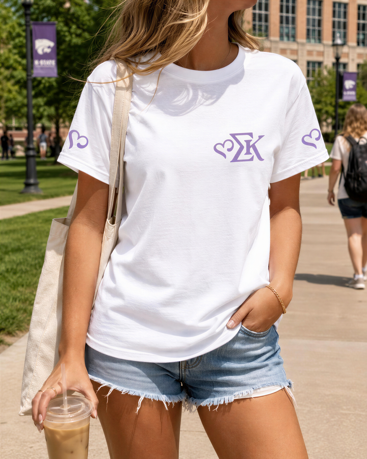 A woman wearing a white t-shirt with a purple logo, denim shorts, and carrying a drink is standing on a sidewalk.