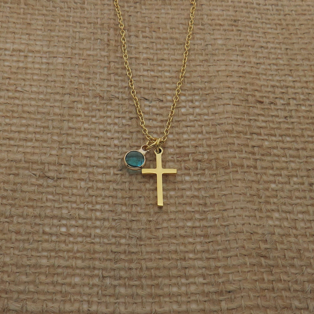 A gold cross necklace with a blue gemstone is displayed against a woven fabric background.