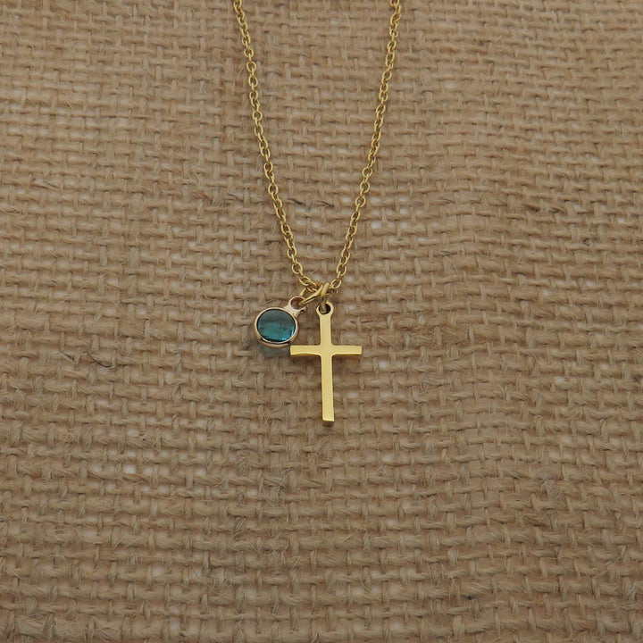 A gold cross necklace with a blue gemstone is displayed against a woven fabric background.