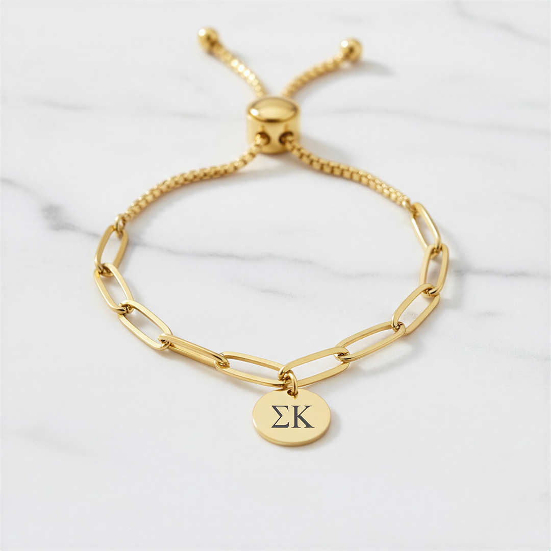 A gold-colored adjustable bracelet with a charm featuring the Greek letter sigma (Σ) on it, placed on a marble surface.
