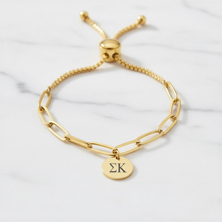 A gold-colored adjustable bracelet with a charm featuring the Greek letter sigma (Σ) on it, placed on a marble surface.