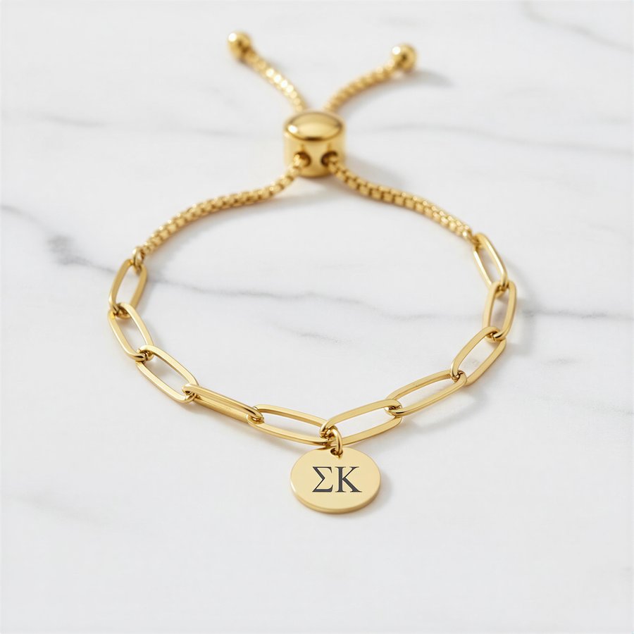 A gold-colored adjustable bracelet with a charm featuring the Greek letter sigma (Σ) on it, placed on a marble surface.