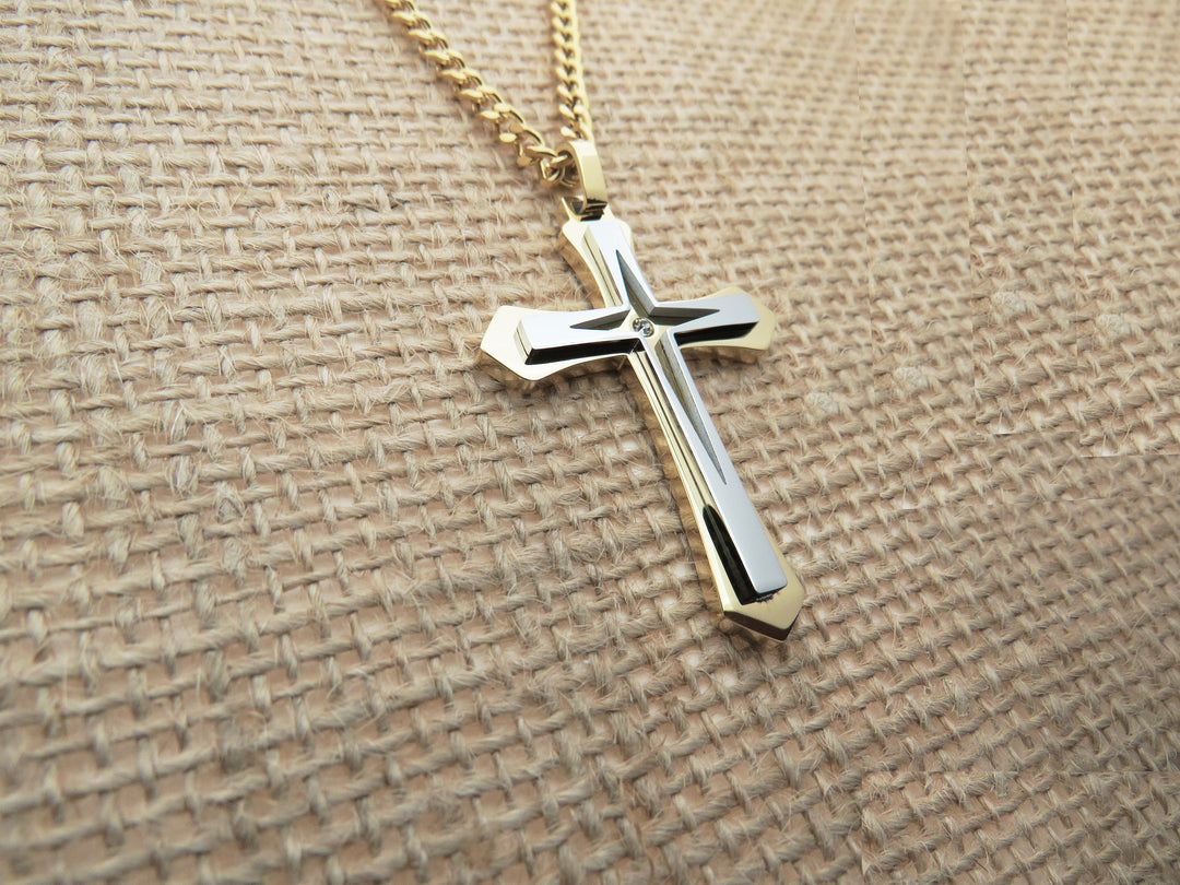 A gold cross pendant with a silver cross design is displayed on a beige woven fabric background.