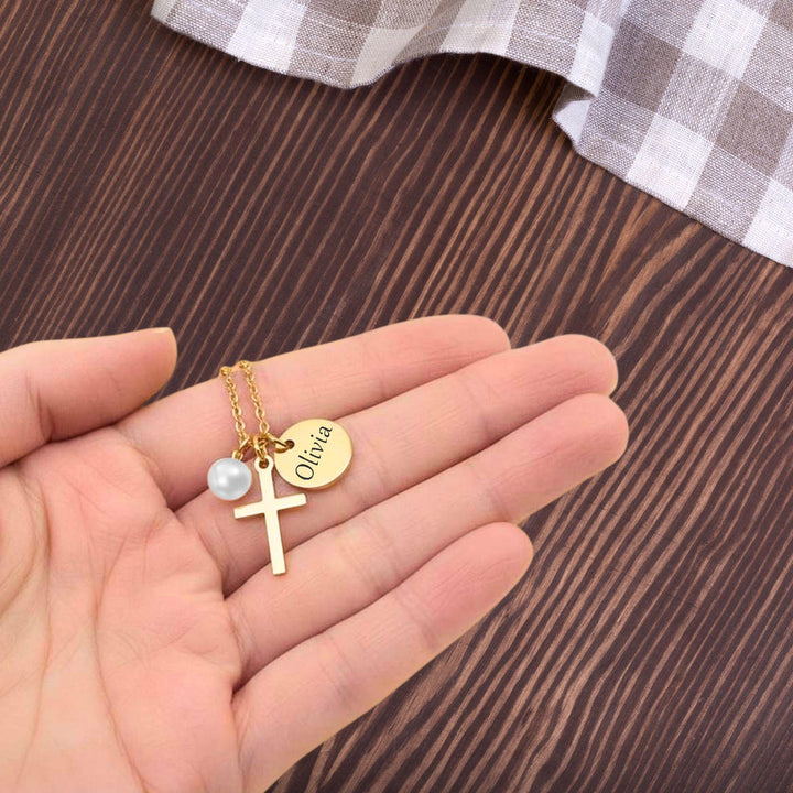 A hand holding a gold cross necklace with a pearl pendant, and a small engraved name tag attached to the necklace.