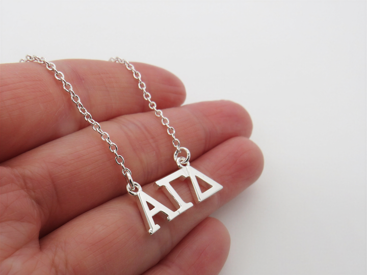 A hand holding a silver necklace with the letters 'ΑΓΑ' on it.