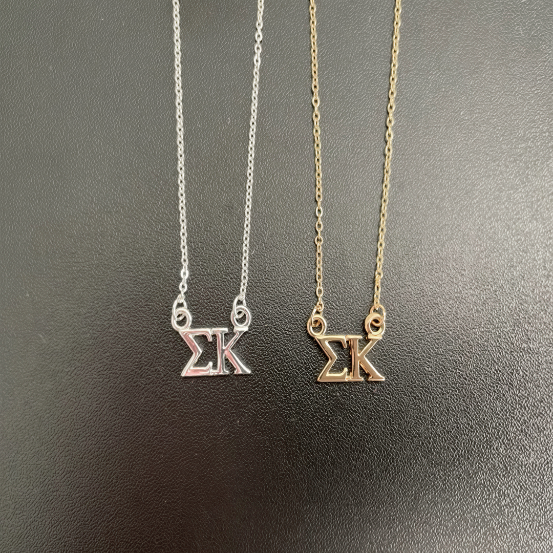 Two necklaces with the letters 'SK' on them, one silver and one gold, are displayed against a dark background.