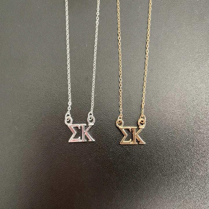 Two necklaces with the letters 'SK' on them, one silver and one gold, are displayed against a dark background.