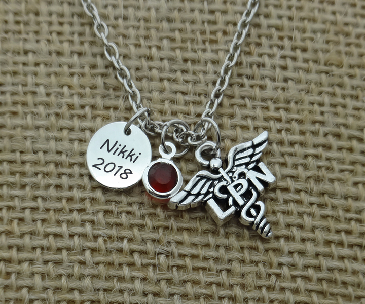 A silver necklace with a charm pendant, featuring a medical symbol and a red gemstone. The charm has the name "Nikki 2018" engraved on it.