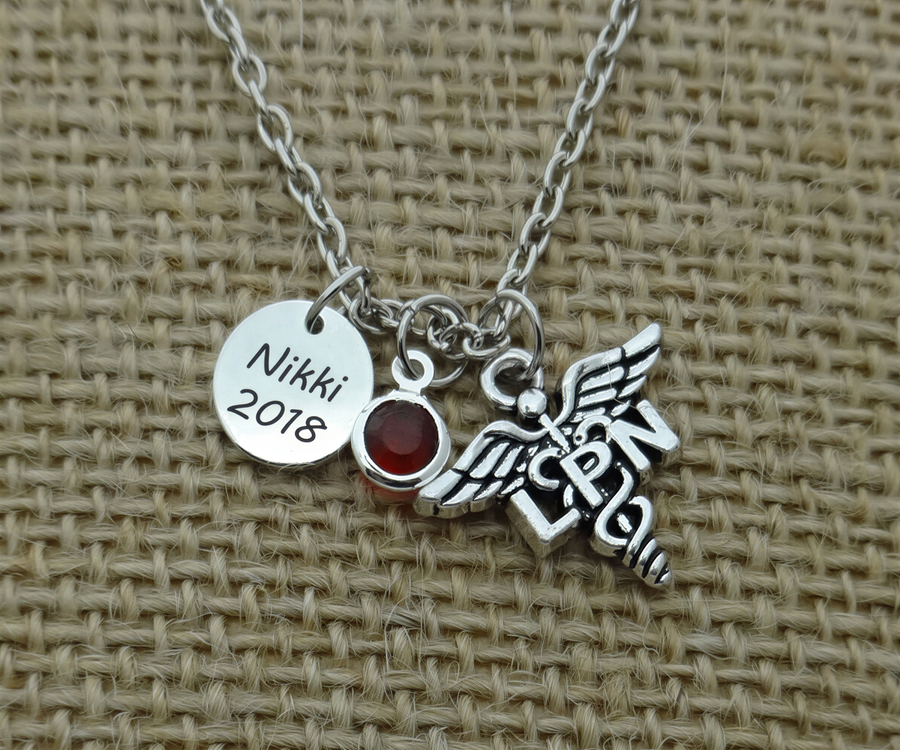 A silver necklace with a charm pendant, featuring a medical symbol and a red gemstone. The charm has the name "Nikki 2018" engraved on it.