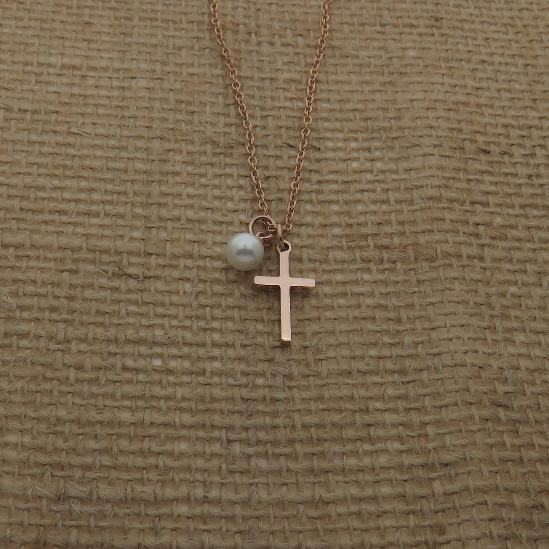 A delicate gold-colored cross pendant with a pearl charm is displayed against a textured beige background.