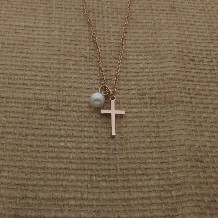 A delicate gold-colored cross pendant with a pearl charm is displayed against a textured beige background.