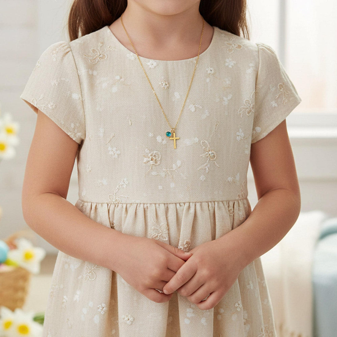 A young girl wearing a beige dress with a floral pattern and a cross necklace stands in a room with a window in the background.
