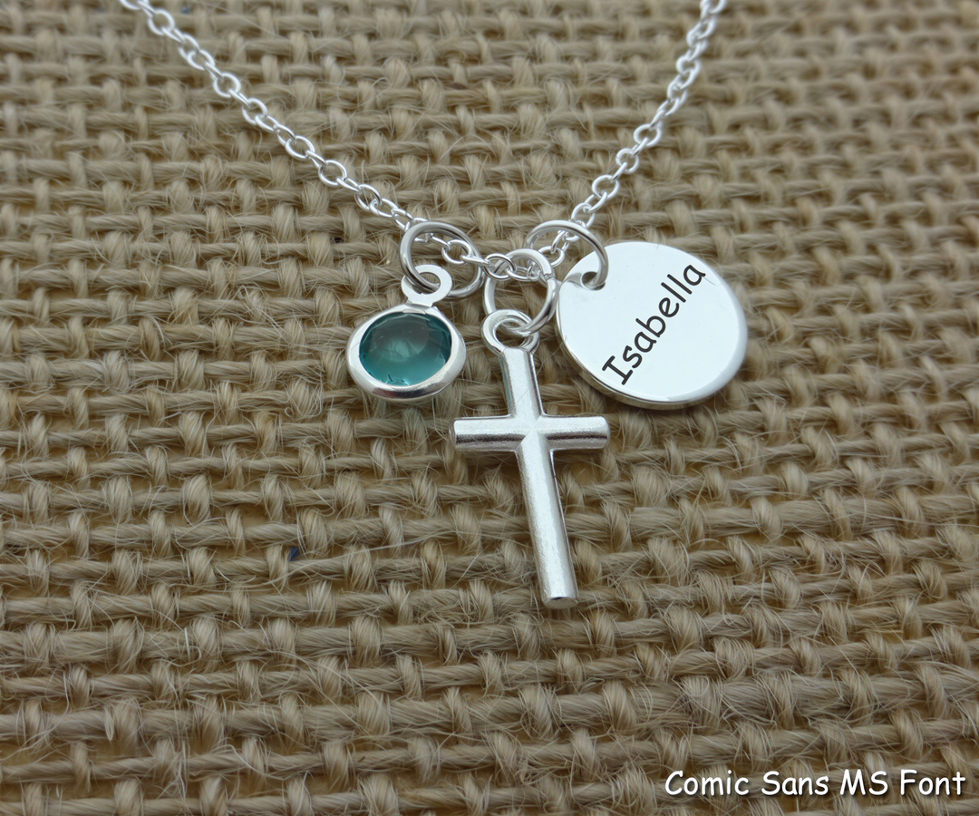 A silver cross necklace with a round charm and a name tag attached to it, resting on a woven surface.