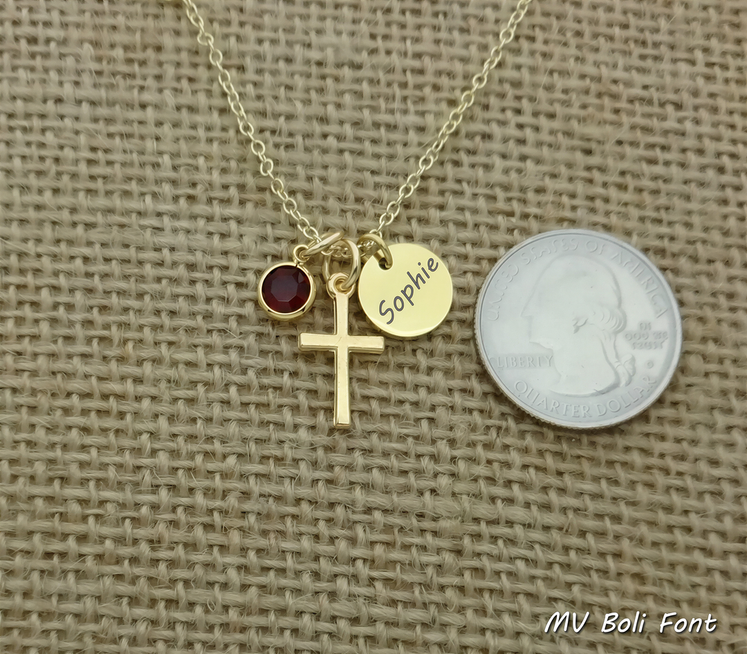 A gold cross necklace with a red gemstone and a small gold charm with the name "Sophie" engraved on it, placed next to a United States quarter.