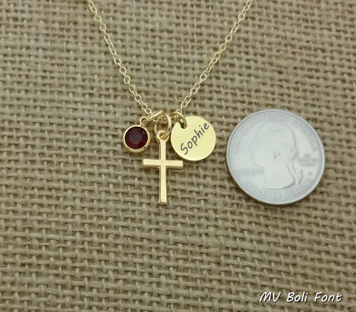A gold cross necklace with a red gemstone and a small gold charm with the name "Sophie" engraved on it, placed next to a United States quarter.