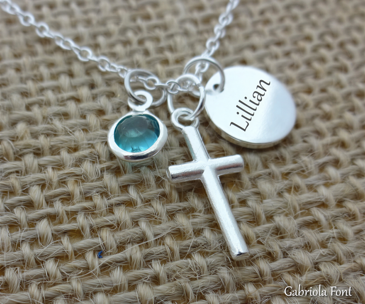 A silver cross pendant with a blue gemstone and a round charm with the name "Lillian" engraved on it, all resting on a woven fabric surface.
