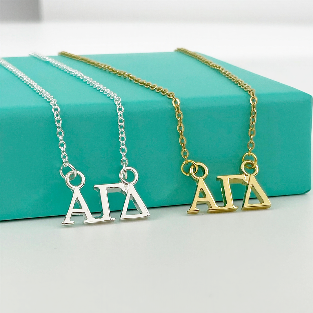 Two necklaces with the letters "ΑΓΑ" on them, one in silver and one in gold, are displayed on a turquoise surface.