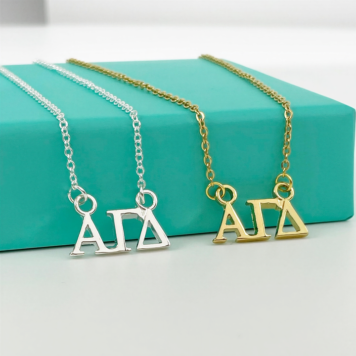 Two necklaces with the letters "ΑΓΑ" on them, one in silver and one in gold, are displayed on a turquoise surface.