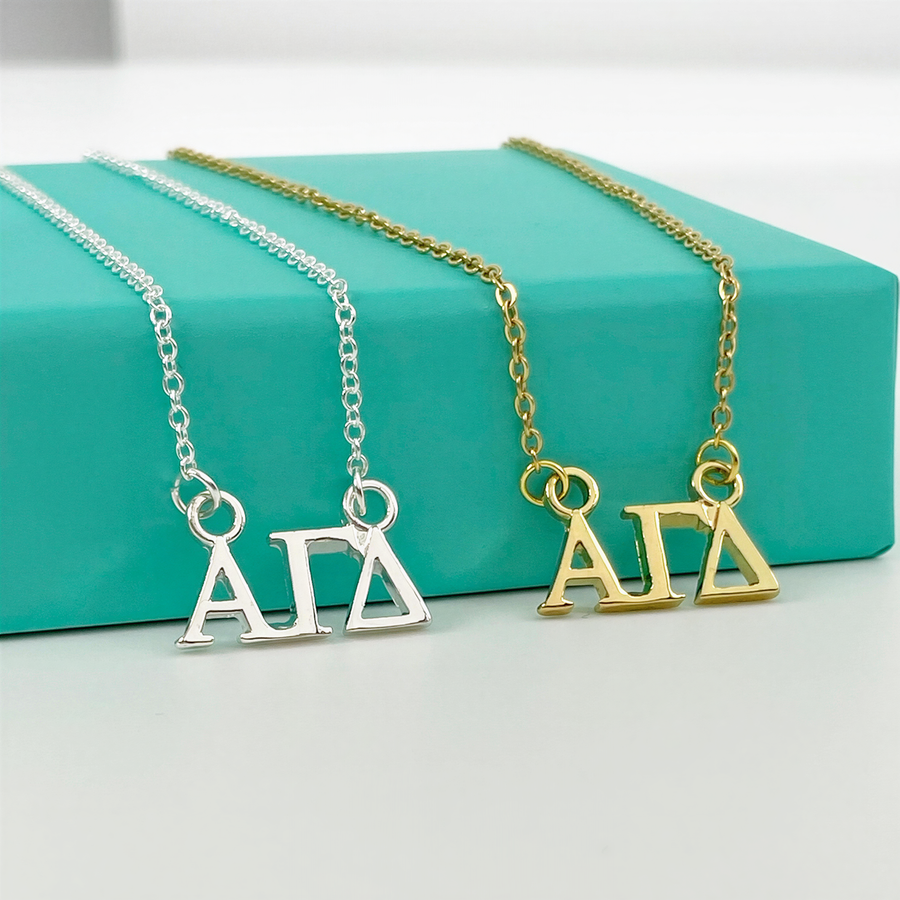 Two necklaces with the letters "ΑΓΑ" on them, one in silver and one in gold, are displayed on a turquoise surface.